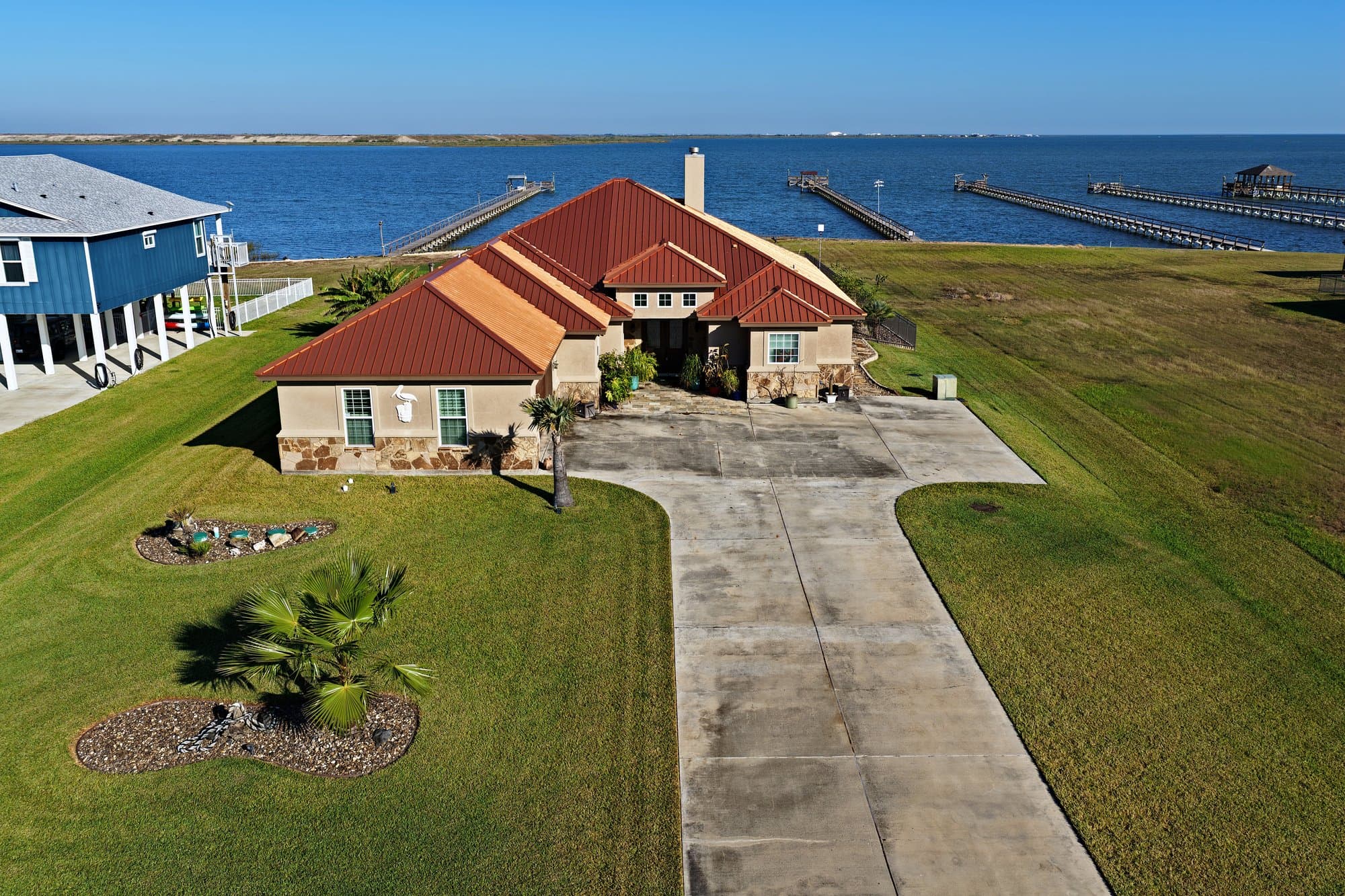 Waterfront house with a red metal roof and long driveway overlooking a bay with piers.