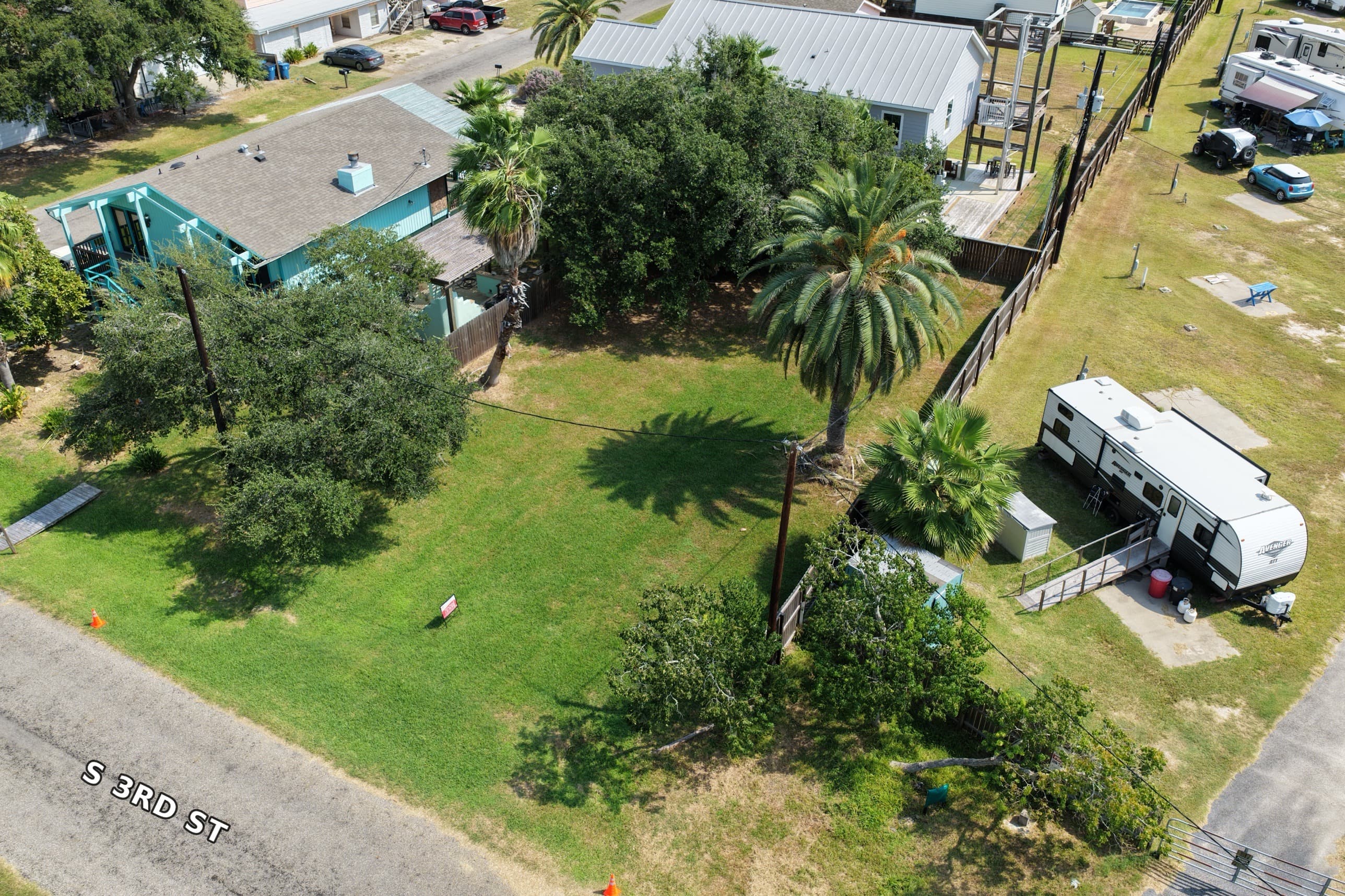 Aerial view of a green vacant lot with palm trees, houses, and a travel trailer.