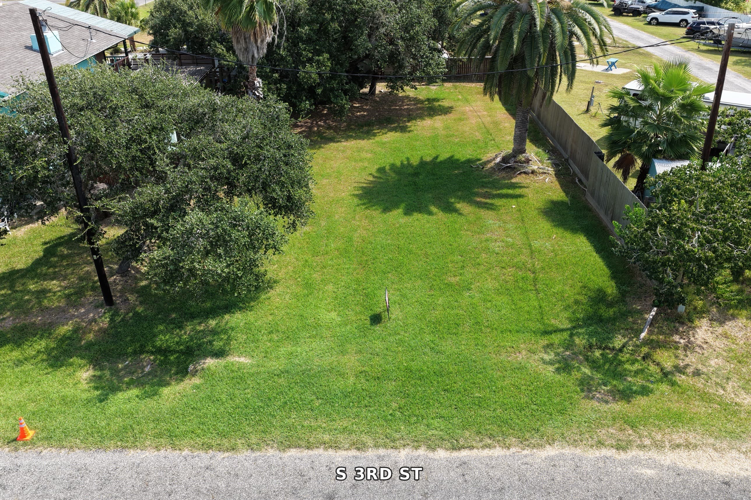 Vacant green lot on S 3rd St with palm trees and a wooden fence.
