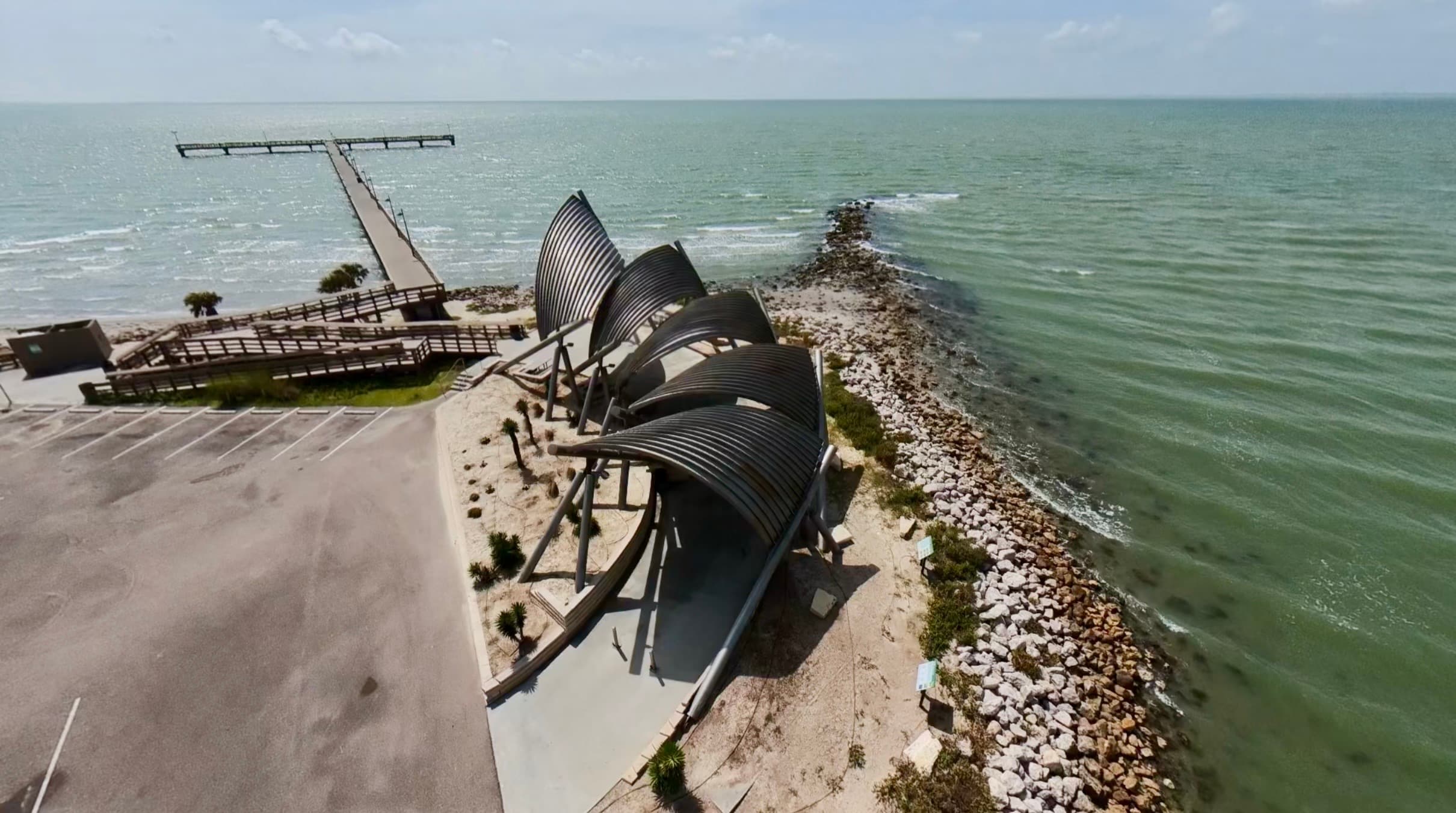 Aerial view of a modern shell-shaped pavilion and long pier along a rocky coastline.