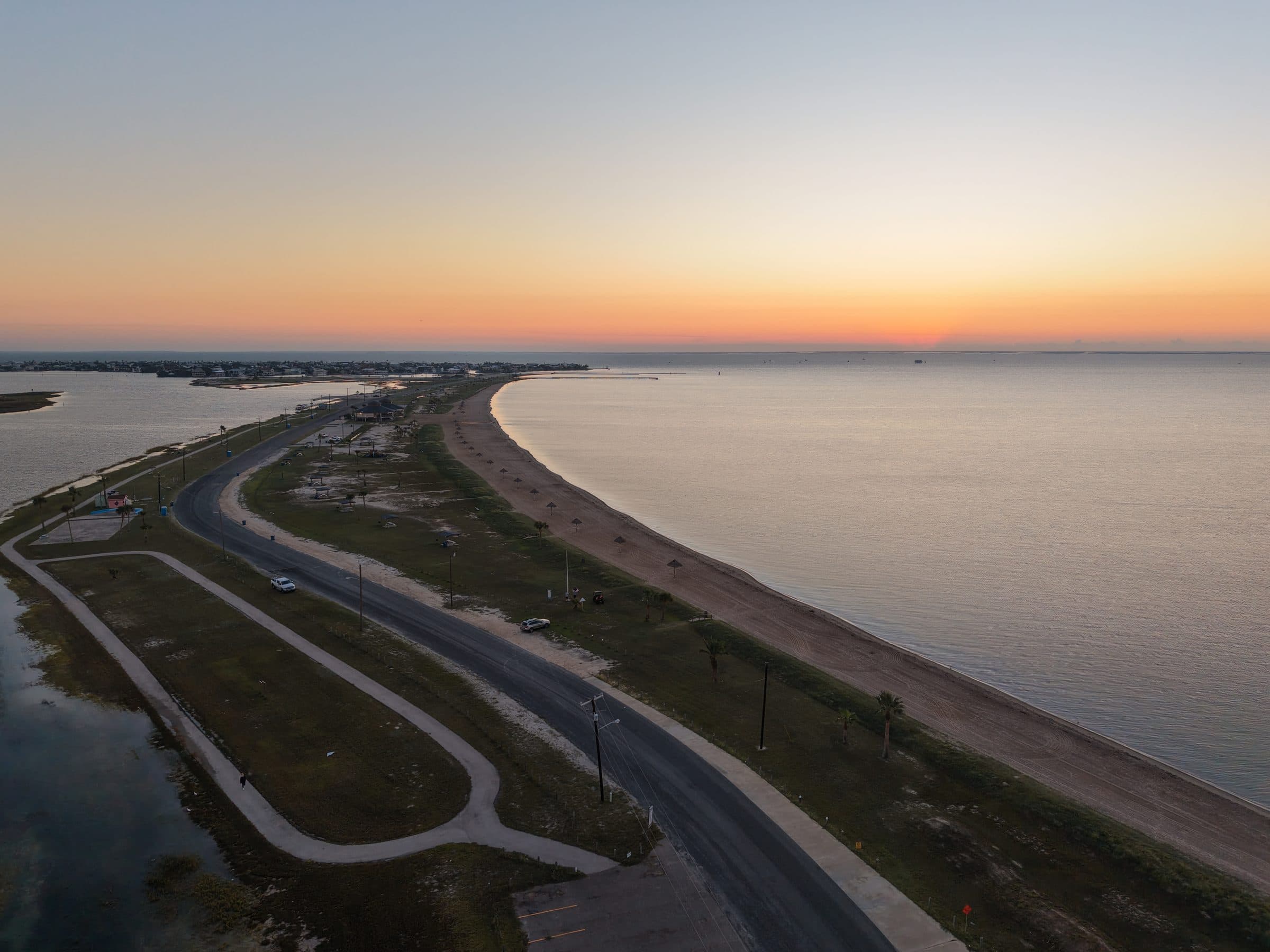 Aerial view of a coastal road and curved beach during a vibrant orange sunset.