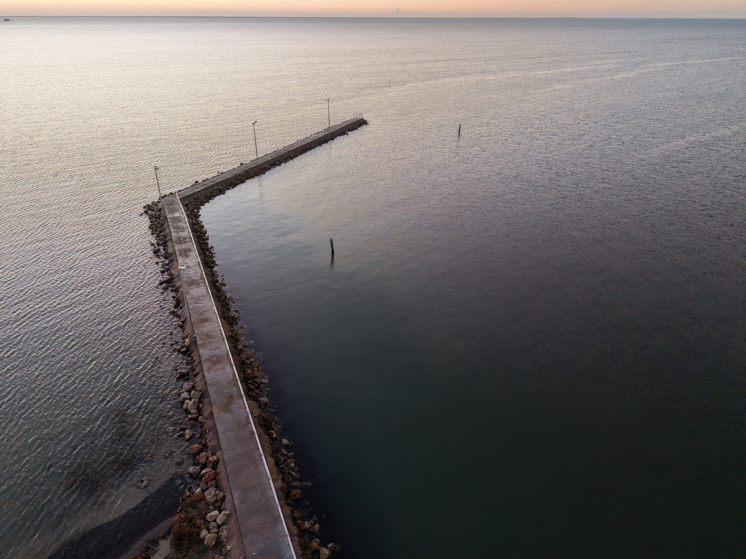 Aerial view of a long, bent concrete pier stretching into a calm, expansive sea.