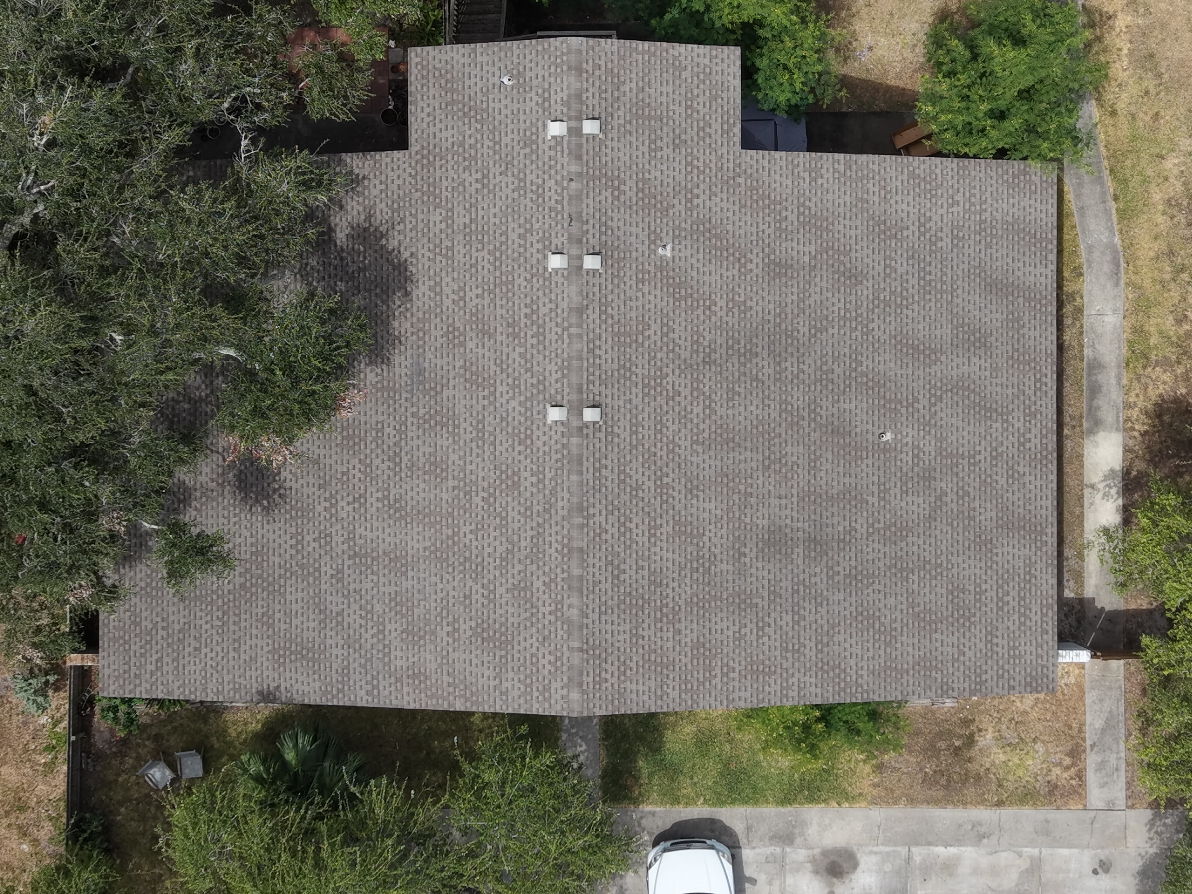 Aerial view of a gray asphalt shingle roof with vents, surrounded by green trees.