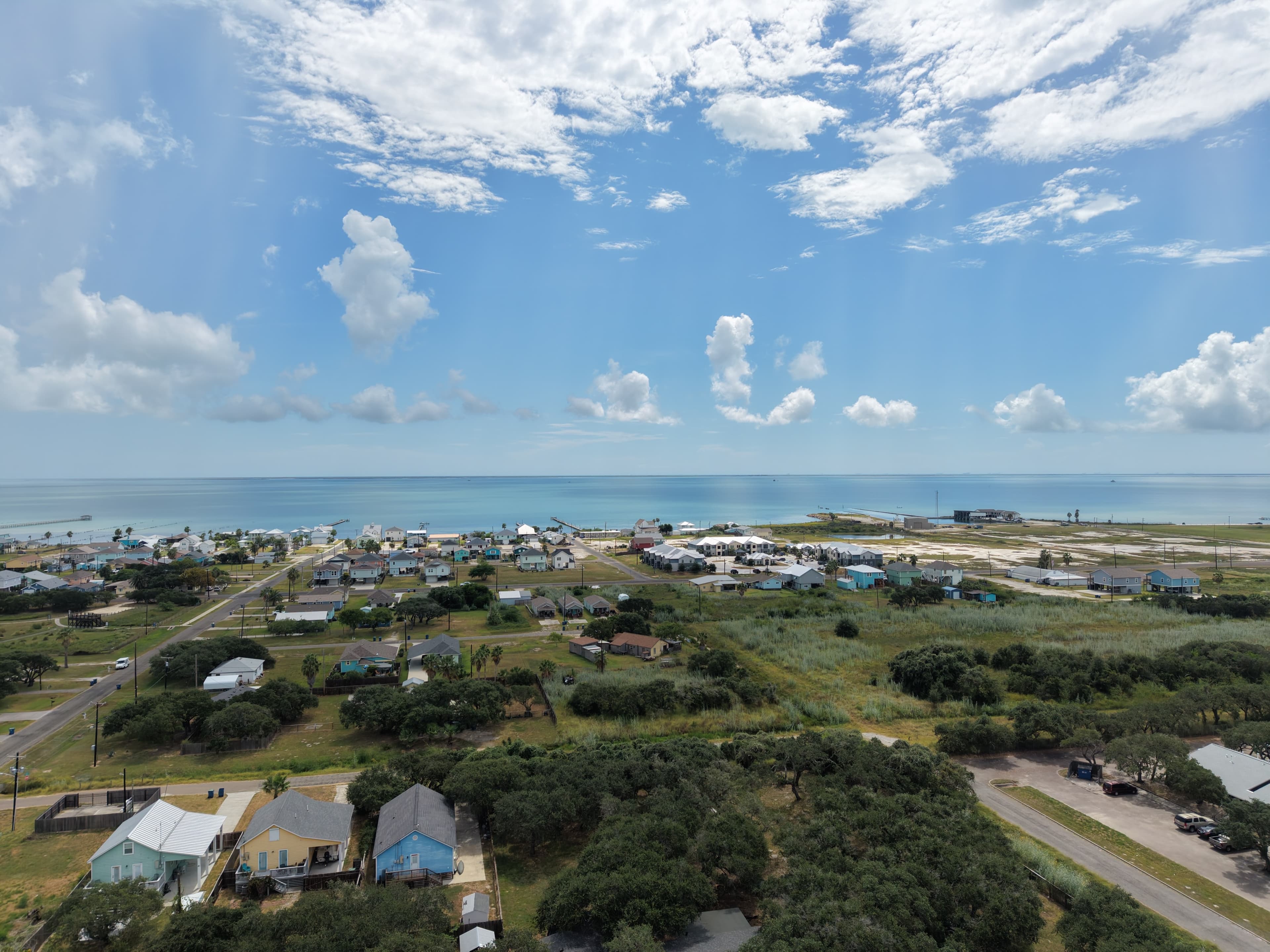 Colorful coastal houses and green trees bordering a calm blue ocean under a bright sky.