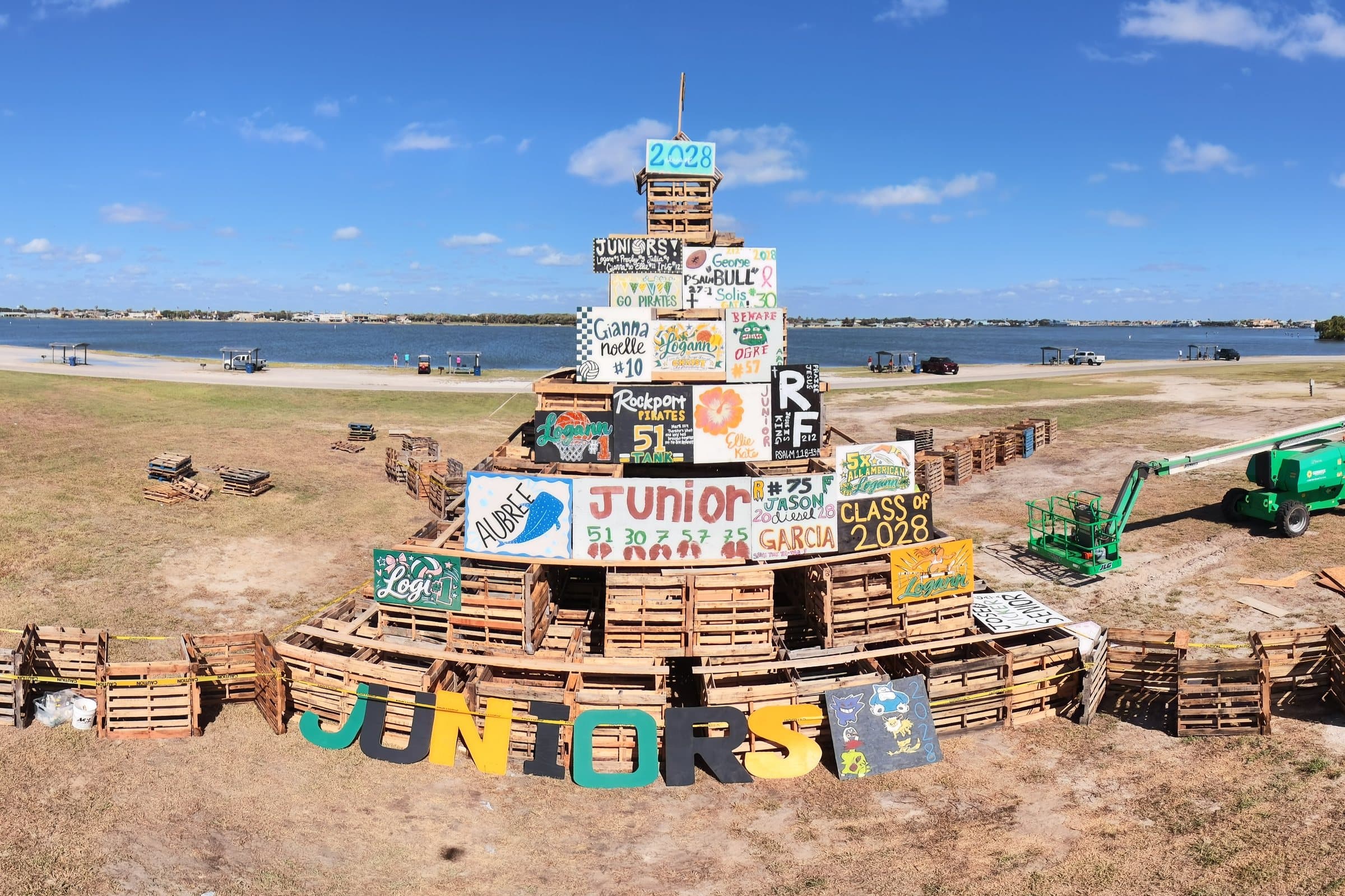 Tiered wooden pallet structure decorated with colorful school spirit signs in a field near water.